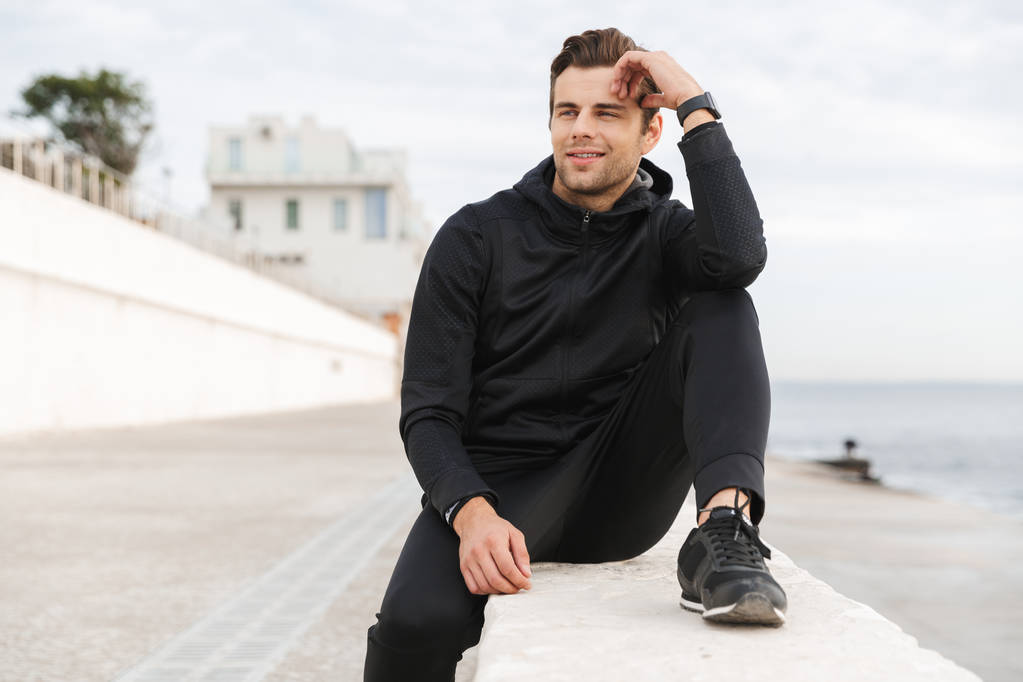 Afbeelding van tevreden sportieve man 30s in zwarte sportkleding zittend op de promenade aan zee - Foto, afbeelding