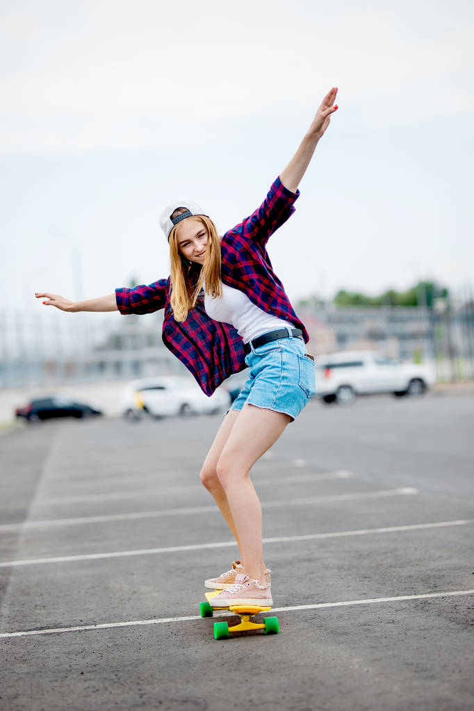 A beautiful blond girl wearing checkered shirt, cap and denim shorts is longboarding while stretching out her hands. Sport and cool style. - Photo, Image
