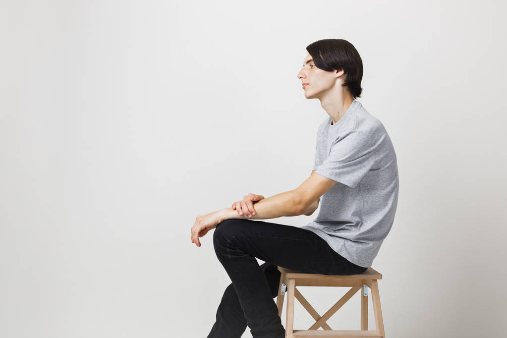 Sakin ve rahat genç ince siyah saçlı bir adam beyaz arka plan karşı sandalyeye oturan gri t-shirt giyen mavi gözlü. Yan görünüm - Fotoğraf, Görsel