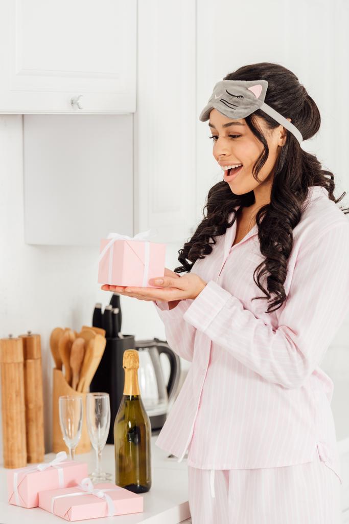 hermosa chica afroamericana excitada en la máscara de dormir sosteniendo caja de regalo rosa en la cocina
 - Foto, Imagen
