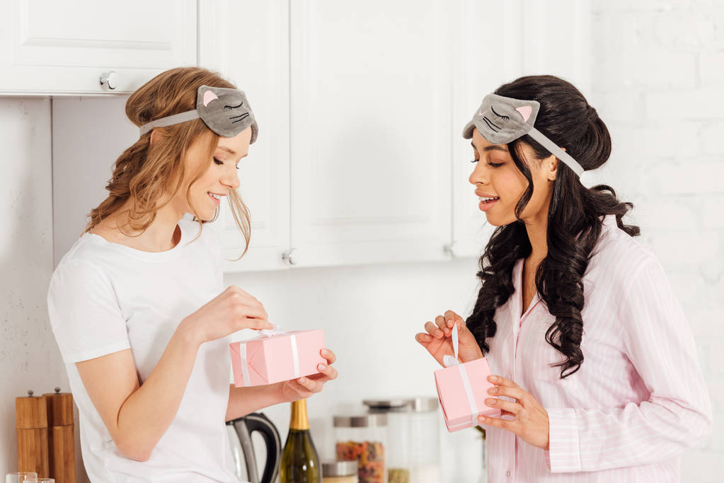 beautiful multicultural girls in sleeping masks and pajamas opening gift boxes in kitchen - Photo, Image