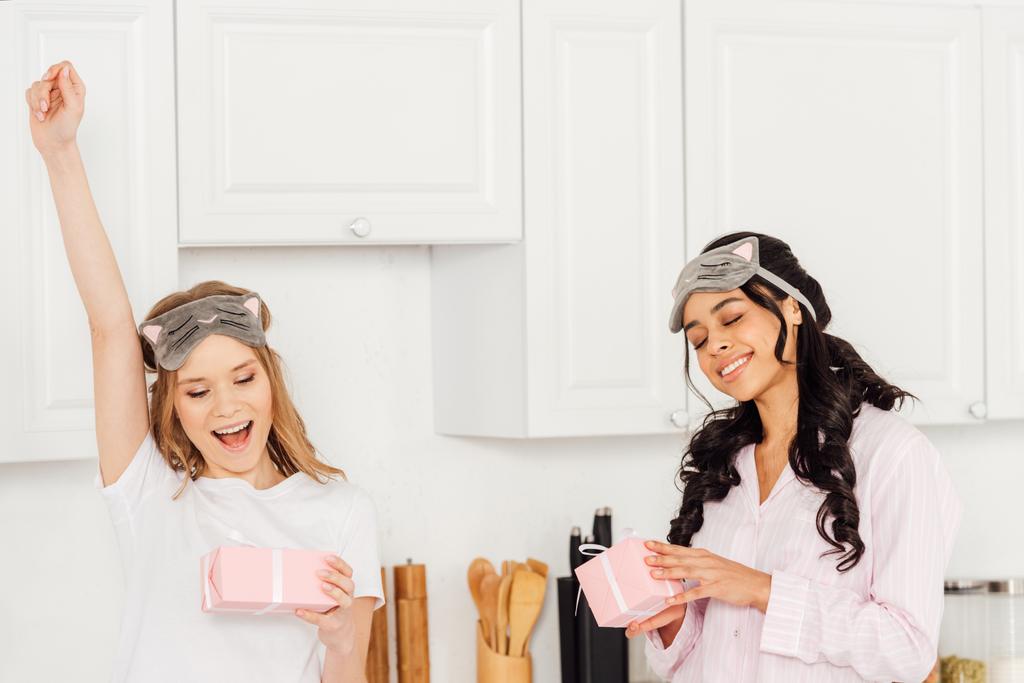 beautiful happy multicultural girls in sleeping masks and pajamas with gift boxes in kitchen - Photo, Image