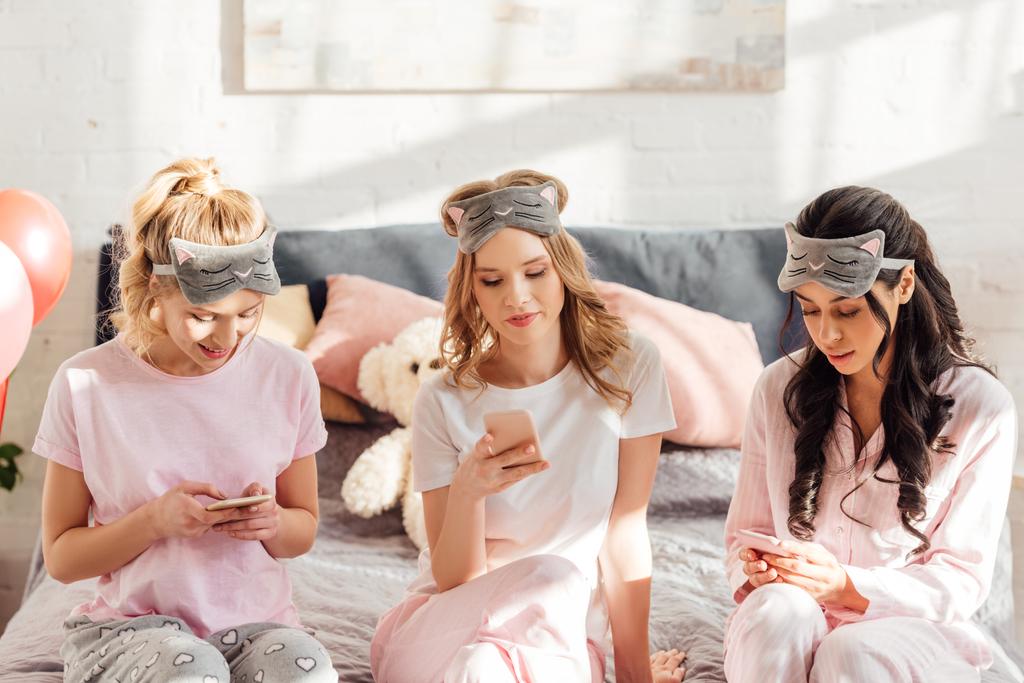 красивые мультикультурные девушки в спальных масках сидят на кровати и используют смартфоны во время пижамной вечеринки
 - Фото, изображение