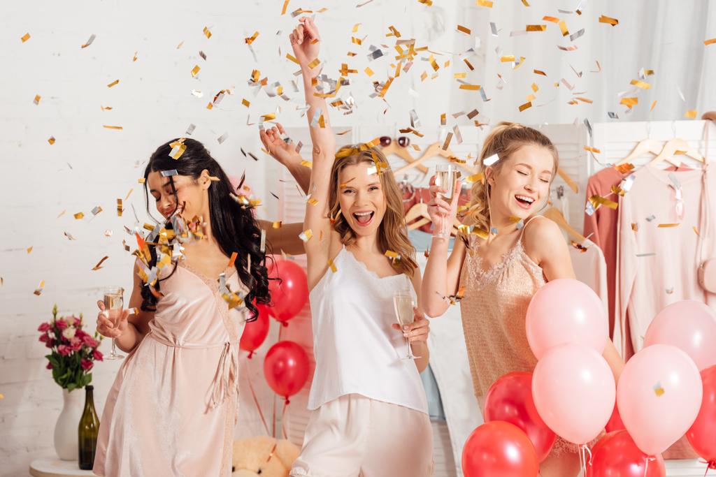 beautiful cheerful multicultural girls holding champagne glasses and celebrating under falling confetti during pajama party - Photo, Image