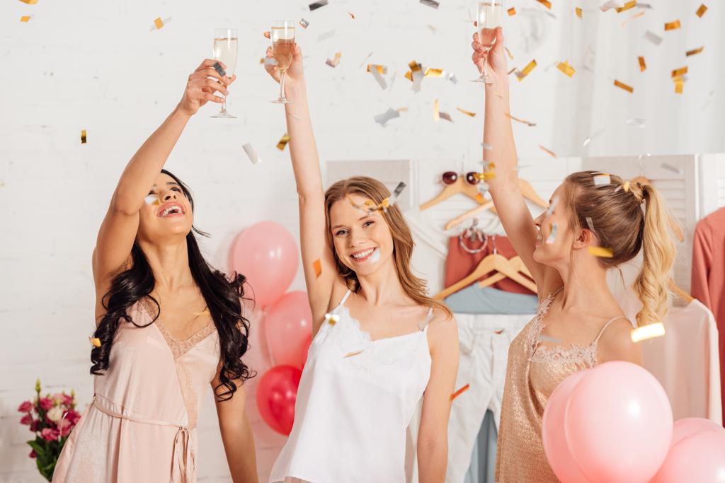 piękne dziewczyny wielokulturowym opiekania z kieliszki do szampana i świętuje pod spadające konfetti w trakcie piżamy strona - Zdjęcie, obraz