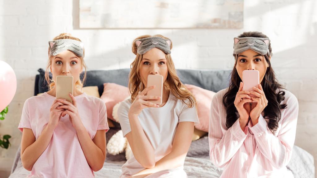 krásné multikulturní dívky v spící masky sedí na posteli a krycí ústa s smartphone během pyžamový večírek - Fotografie, Obrázek