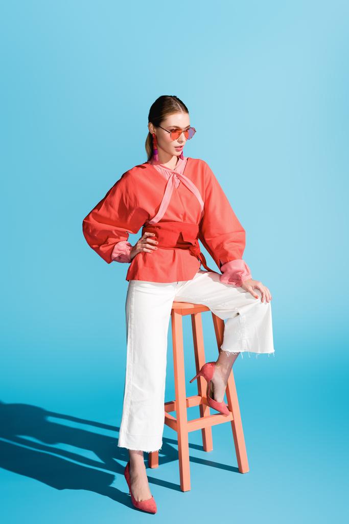 attractive woman posing in living coral clothing and sunglasses on stool on blue - Photo, Image