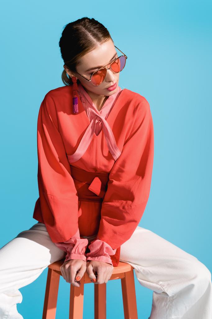 stylish woman in trendy living coral clothing and sunglasses posing on stool isolated on blue - Photo, Image