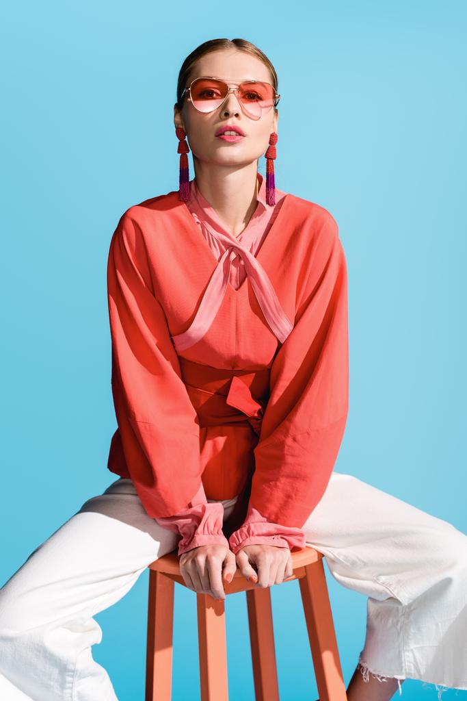 модная модель в модной живой коралловой одежде и солнцезащитных очках, позирующих на стуле, изолированном от бирюзы
 - Фото, изображение