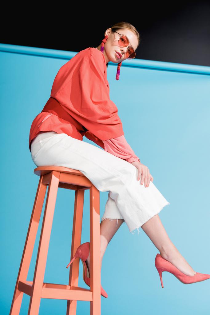 beautiful woman in trendy living coral clothing and sunglasses posing on stool on blue - Photo, Image