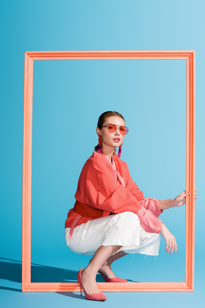 fille à la mode dans des vêtements de corail vivant et des lunettes de soleil posant avec un grand cadre sur bleu
 - Photo, image