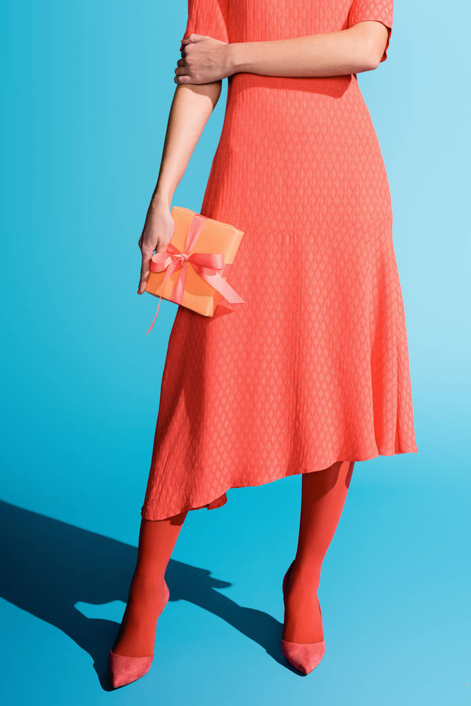 bijgesneden weergave van stijlvolle meisje in levende koraal jurk bedrijf gift box op blauw - Foto, afbeelding