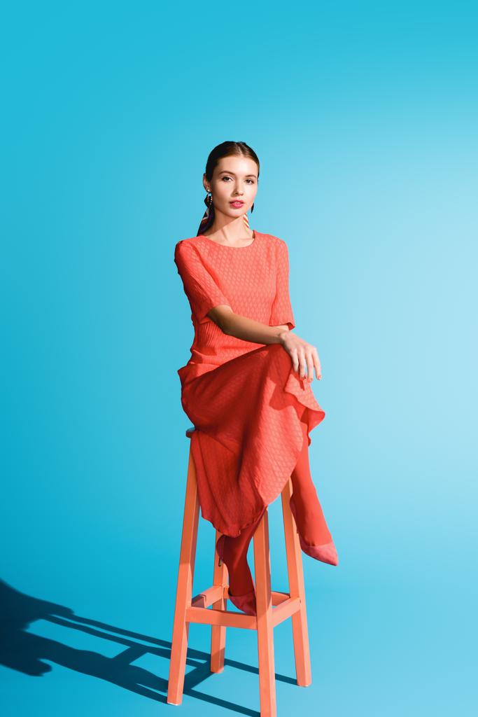 стильная девушка в модном живом коралловом платье позирует на стуле на голубом
 - Фото, изображение