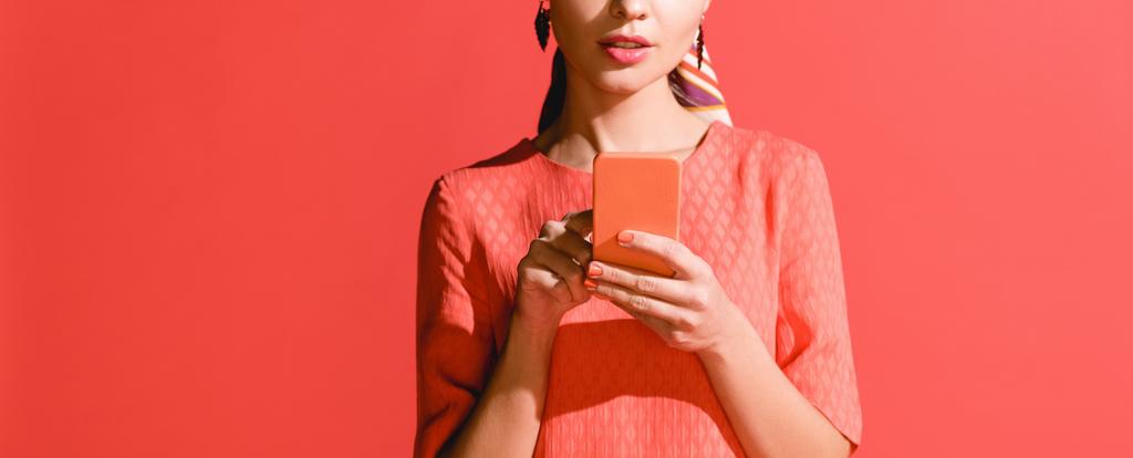 vue recadrée de fille en robe de corail vivant en utilisant smartphone sur rouge. Couleur Pantone de l'année 2019 concept
 - Photo, image