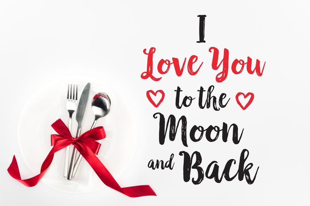 erhöhter Blick auf Gabel, Messer und Löffel, umwickelt von roter Festschleife auf Teller isoliert auf weißem, Valentinstag-Konzept mit dem Schriftzug "Ich liebe dich zum Mond und zurück" - Foto, Bild