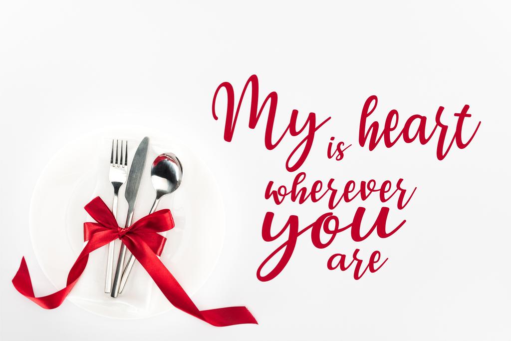 elevated view of fork, knife and spoon wrapped by red festive bow on plate isolated on white, st valentine day concept with "my heart is wherever you are" lettering - Photo, Image