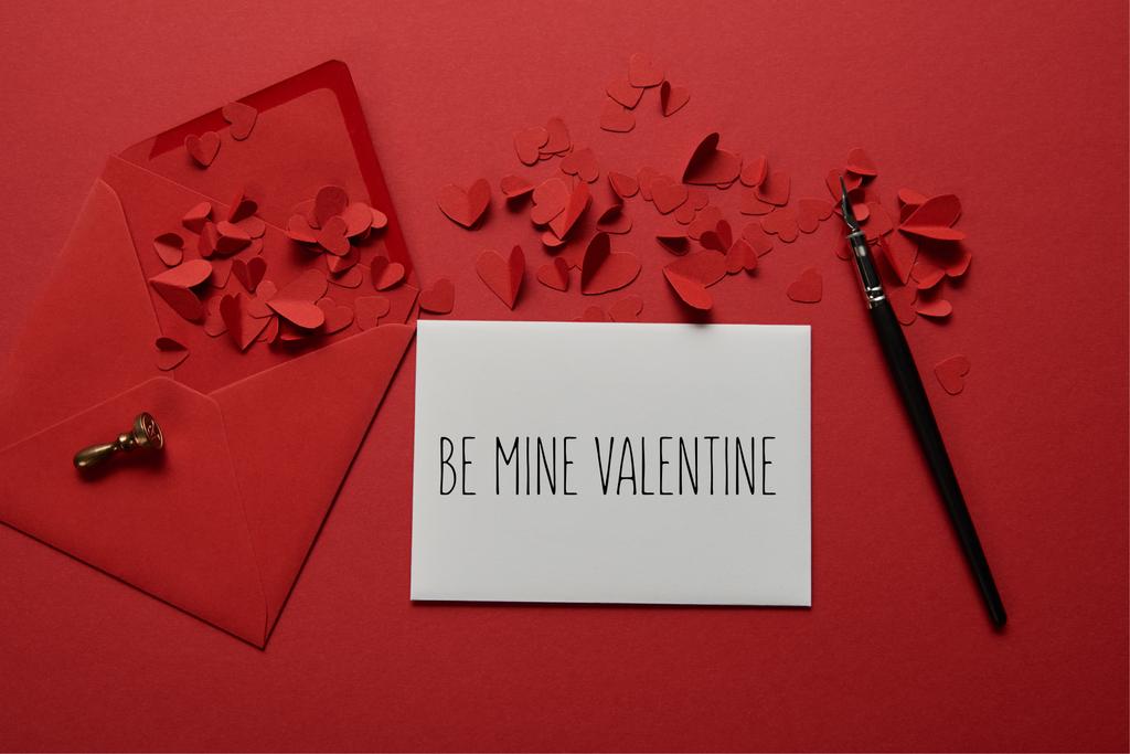 top view of envelope with "be mine valentine" lettering , paper cut hearts and pen on red background - Photo, Image