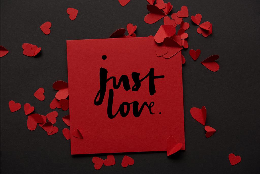 bovenaanzicht van rode wenskaart met "gewoon love" letters en papier knippen harten op zwarte achtergrond - Foto, afbeelding