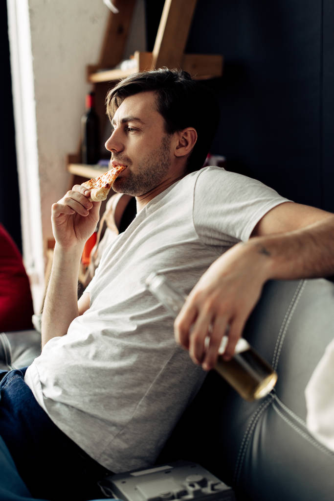 mies syö maukasta pizzaa ja pitää pullon olohuoneessa
 - Valokuva, kuva