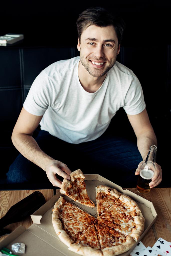 vrolijke man met stukje heerlijke pizza en fles in woonkamer - Foto, afbeelding