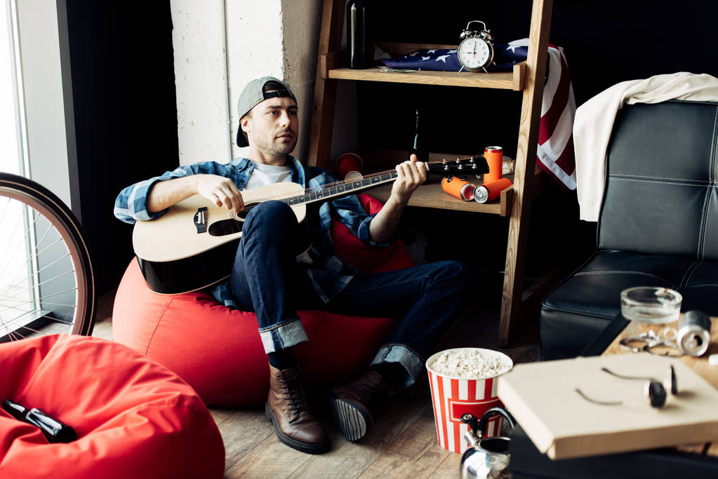 άνδρας στο καπάκι παίζουν ακουστική κιθάρα στο ακατάστατο σαλόνι   - Φωτογραφία, εικόνα