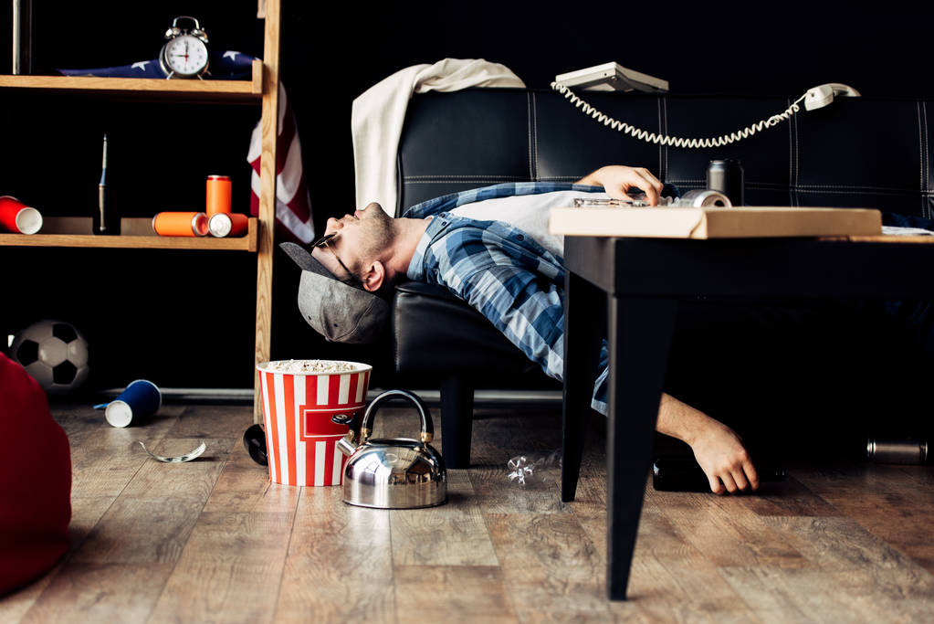 человек в шапке лежит на диване возле коробки попкорна и чайник после вечеринки
 - Фото, изображение