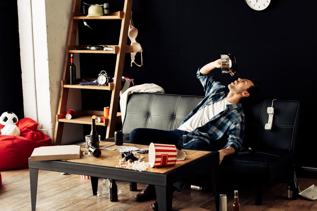 borracho bebiendo de la tetera mientras está sentado en el sofá en la sala de estar desordenada
  - Foto, Imagen