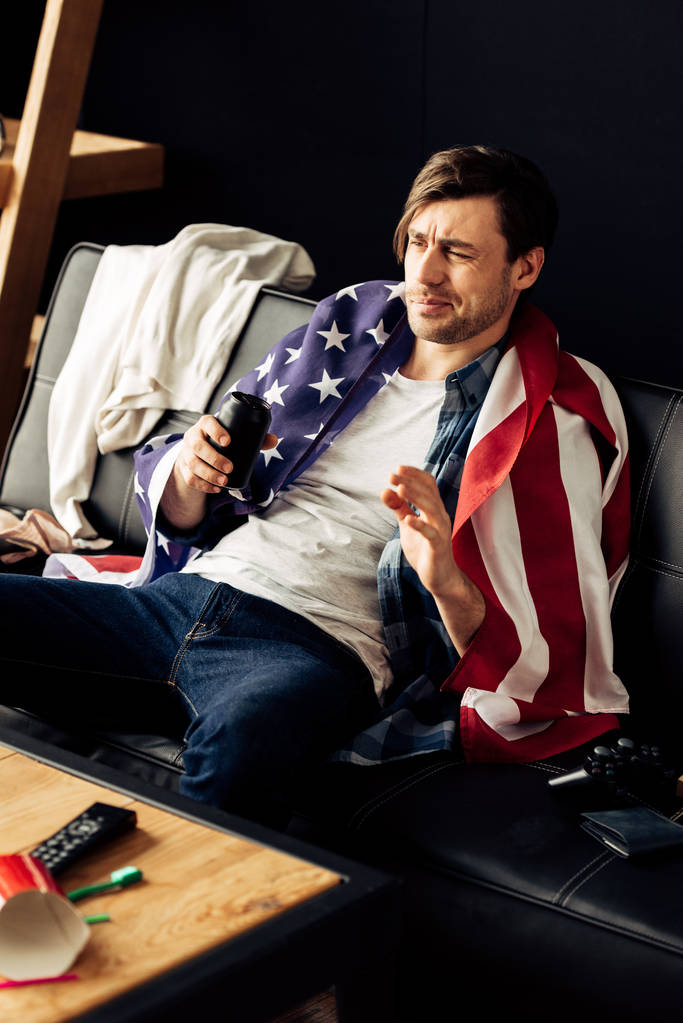 drunk man holding can with beer and sitting with american flag on sofa after party - Photo, Image
