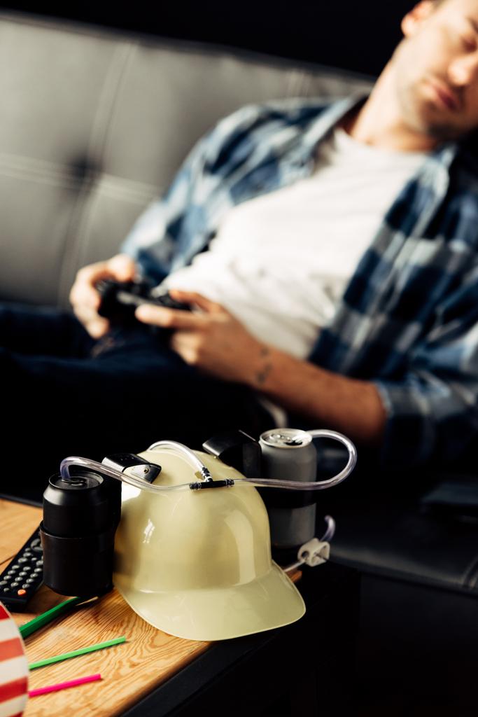 selective focus of beer helmet with man sleeping and holding joystick on background - Photo, Image