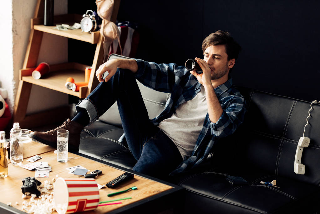 п'яний чоловік сидить на дивані і п'є пиво в безладному квартирі
  - Фото, зображення
