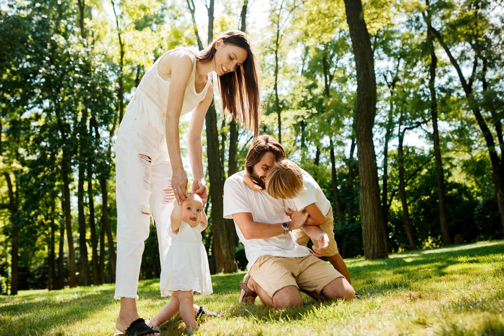 Щасливі молоді темноволосої матері і батька, одягнені в білий одяг, проводять літній день з дітьми на газоні в парку
. - Фото, зображення