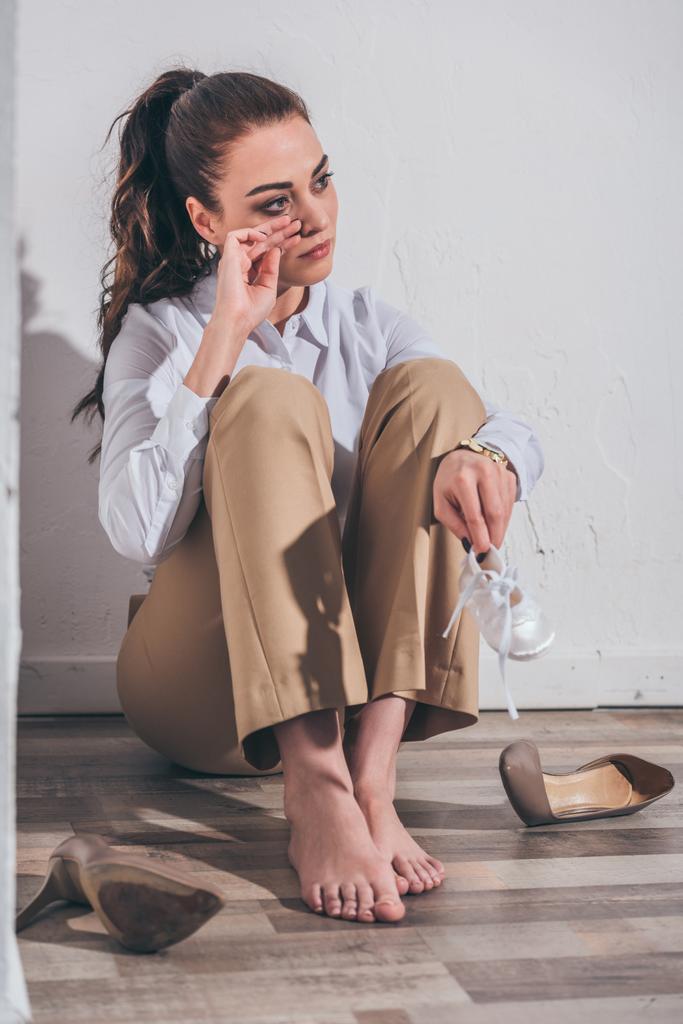 расстроена женщина сидит на полу, плачет и держа детские туфли возле белой стены дома, скорбящее расстройство концепции
 - Фото, изображение