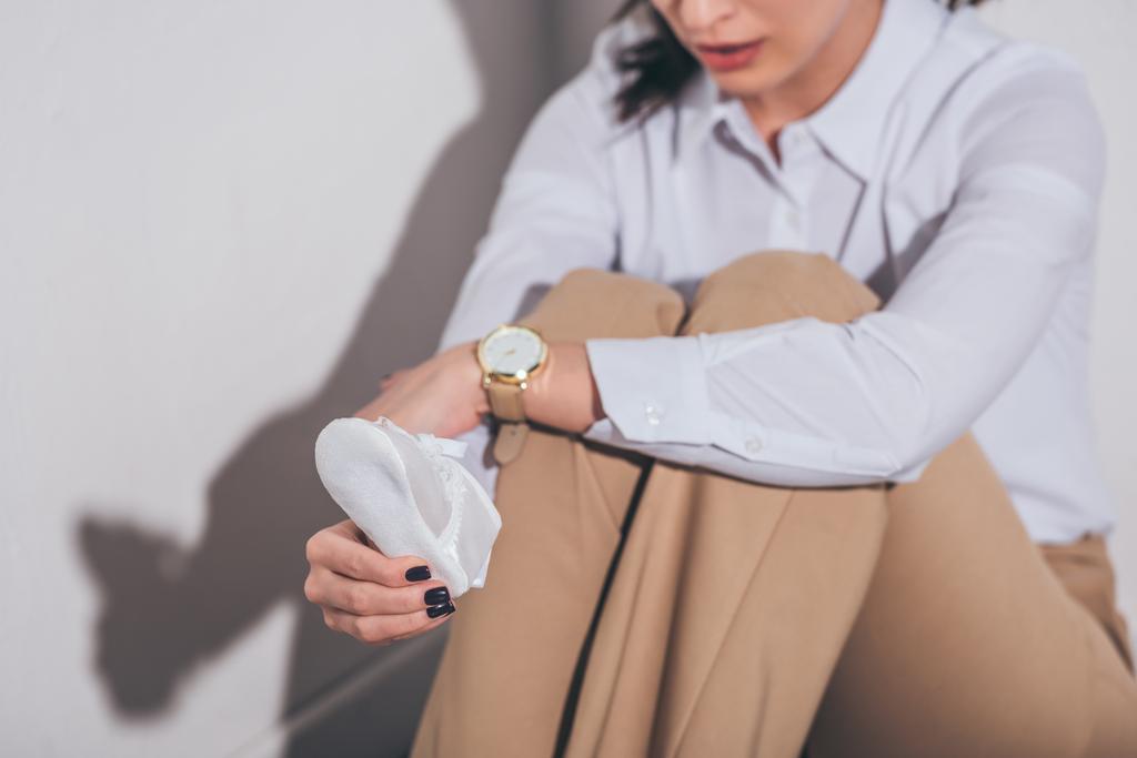 vue recadrée de femme triste en chemisier blanc et pantalon beige assis sur le sol près du mur et tenant chaussure de bébé à la maison, concept de trouble du deuil
 - Photo, image