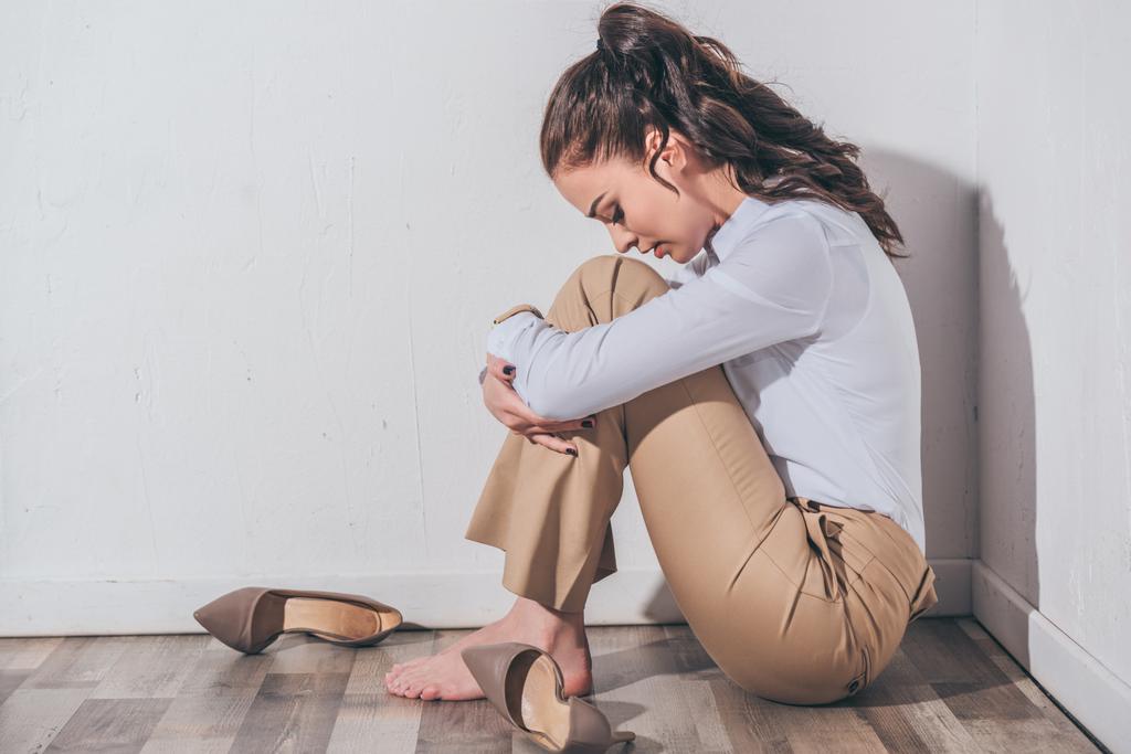 mujer triste en blusa blanca y pantalones beige sentado en el suelo con la cabeza doblada cerca de la pared blanca en casa, concepto de trastorno de duelo
 - Foto, Imagen
