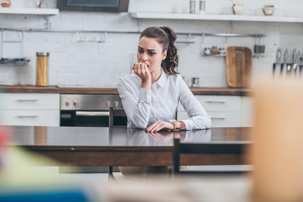zdenerwowany Kobieta w białej bluzce siedzi tabeli ar i bitting paznokcie w kuchni, żałoby koncepcja zaburzenia - Zdjęcie, obraz
