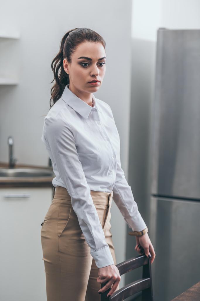расстроена женщина в белой блузке и бежевых брюках, стоящих на стуле на кухне, скорбящее расстройство концепции
 - Фото, изображение