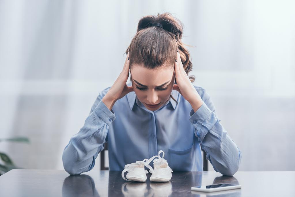 сумна жінка в блакитній блузці, сидячи на дерев'яному столі з смартфоном і дивлячись на біле дитяче взуття вдома, сумна концепція розладу
 - Фото, зображення