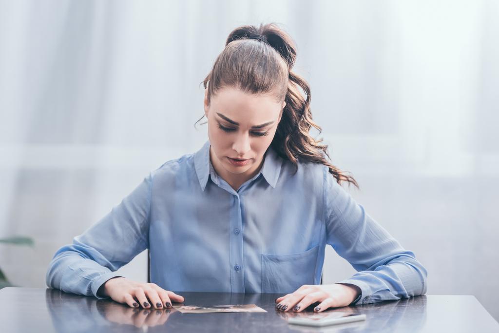 upset woman in blue blouse sitting at wooden table with smartphone and looking at photo in room, grieving disorder concept - Photo, Image