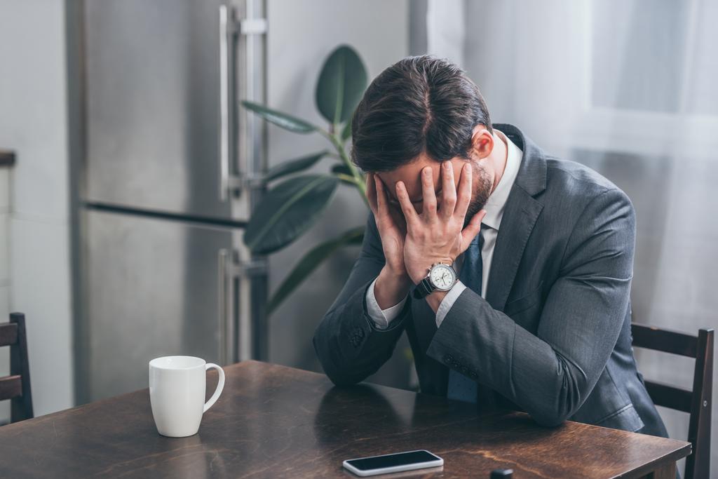 smutný muž v šedém obleku, sedí u stolu s smartphone, bílé víčko a pokrývající obličej rukama doma, truchlící porucha koncept - Fotografie, Obrázek