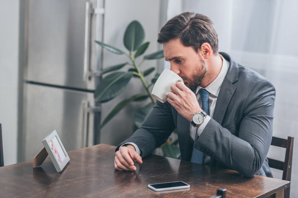 triste hombre en traje gris sentado en la mesa de madera con el teléfono inteligente beber y mirando poto en el marco en casa, concepto de trastorno de duelo
 - Foto, imagen
