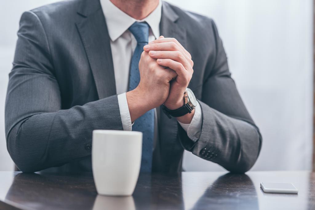 Ausgeschnittene Ansicht eines Mannes im grauen Anzug, der mit weißer Tasse und Smartphone zu Hause am Holztisch sitzt und trauert. - Foto, Bild