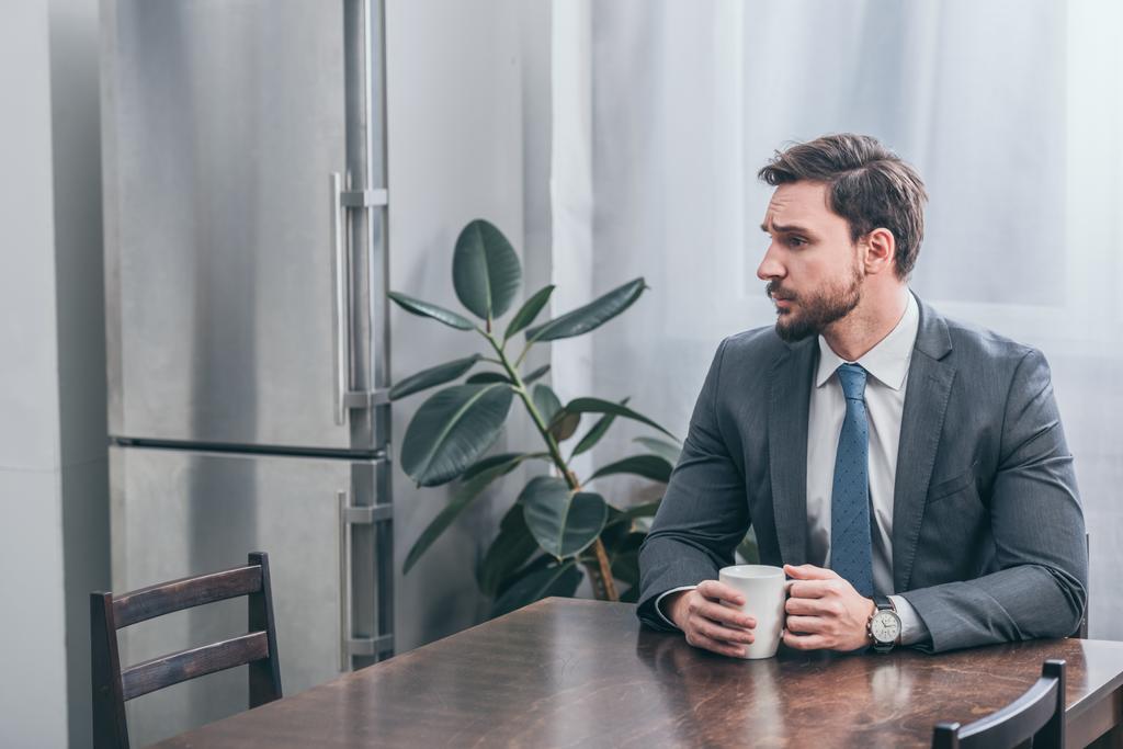 smutný muž v šedém obleku sedí u dřevěného stolu a drží bílé pohár doma, truchlící porucha koncept - Fotografie, Obrázek