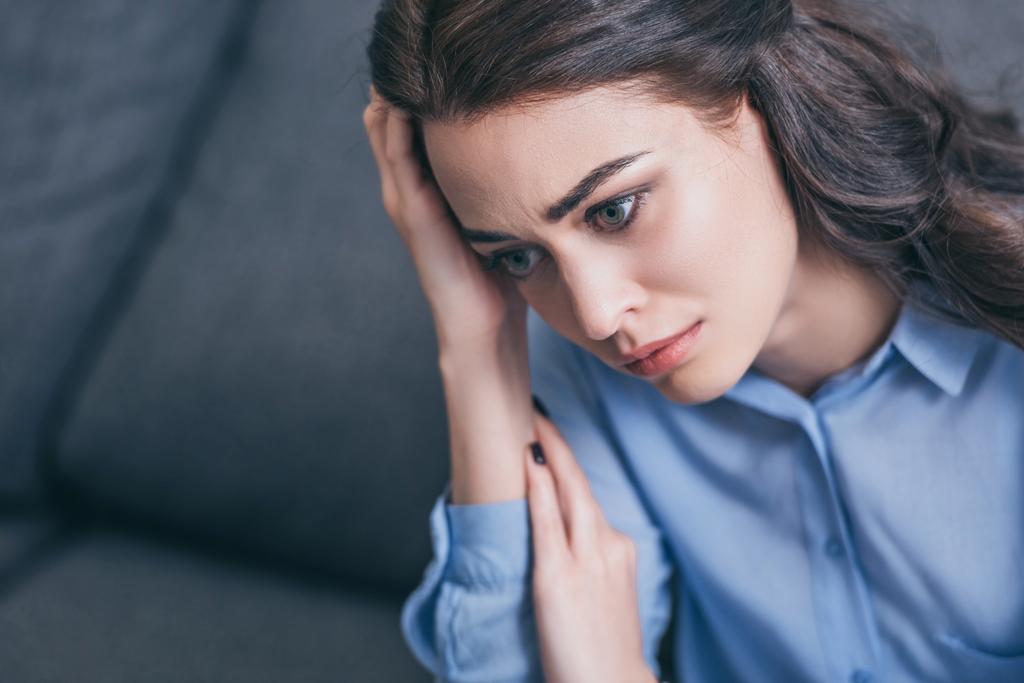 sad woman in blue blouse sitting on grey couch and holding head in room, grieving disorder concept - Photo, Image
