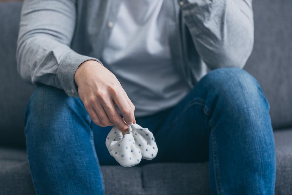 обрізаний вид чоловіка, що сидить на сірому дивані і тримає шкарпетки для немовлят вдома, сувора концепція розладу
 - Фото, зображення