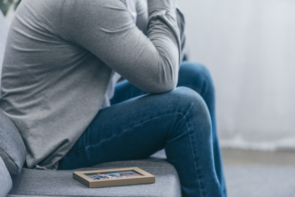 cropped view of man in grey shirt and blue pants sitting on couch with photo frame at home, grieving disorder concept - Photo, Image