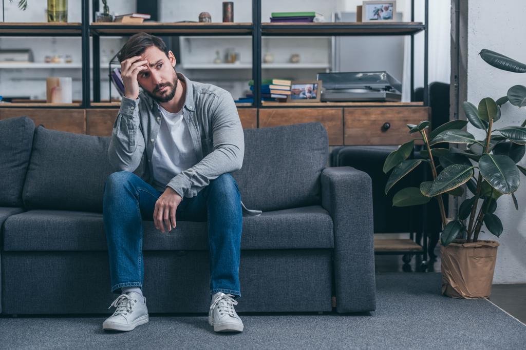 Depressiver Mann sitzt auf Couch, berührt Gesicht und trauert zu Hause - Foto, Bild
