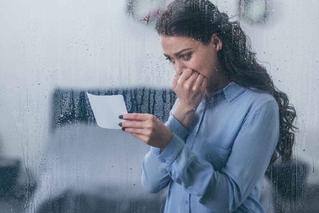 sureva nainen pitelee valokuvaa, peittää suun käsin ja itkee kotona ikkunan läpi sadepisaroilla
 - Valokuva, kuva