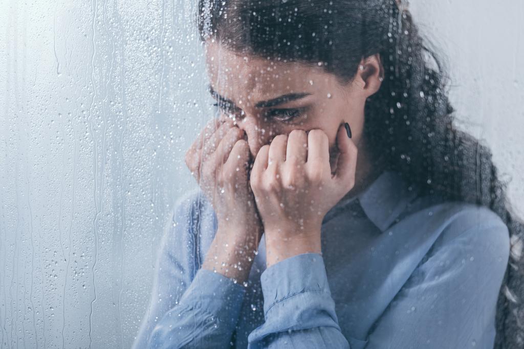 καταθλιπτική γυναίκα που καλύπτουν το πρόσωπο με τα χέρια και κλαίγοντας στο σπίτι μέσα από το παράθυρο με τις σταγόνες της βροχής - Φωτογραφία, εικόνα