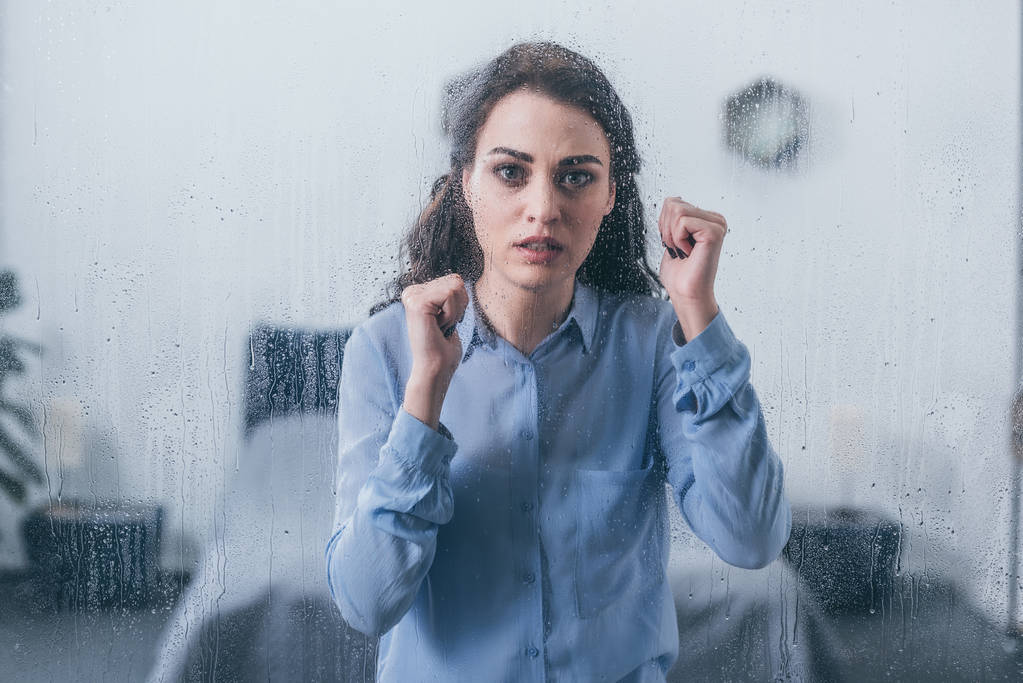 beautiful sad woman with clenched fists looking at camera through window with raindrops - Photo, Image