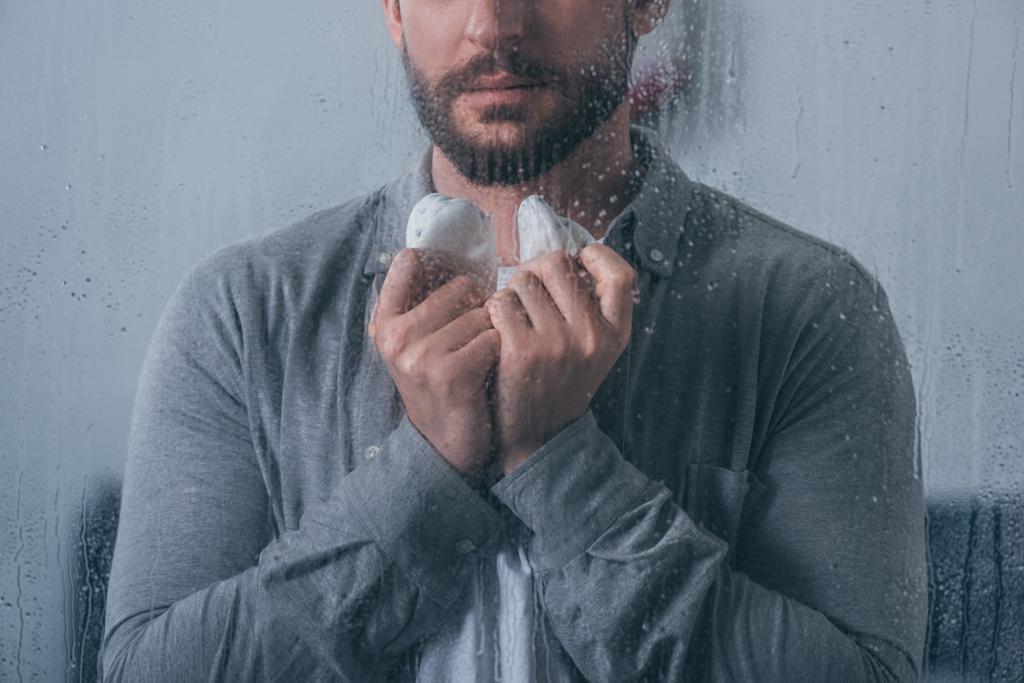 cropped view of man holding baby shoes through window with raindrops - Photo, Image
